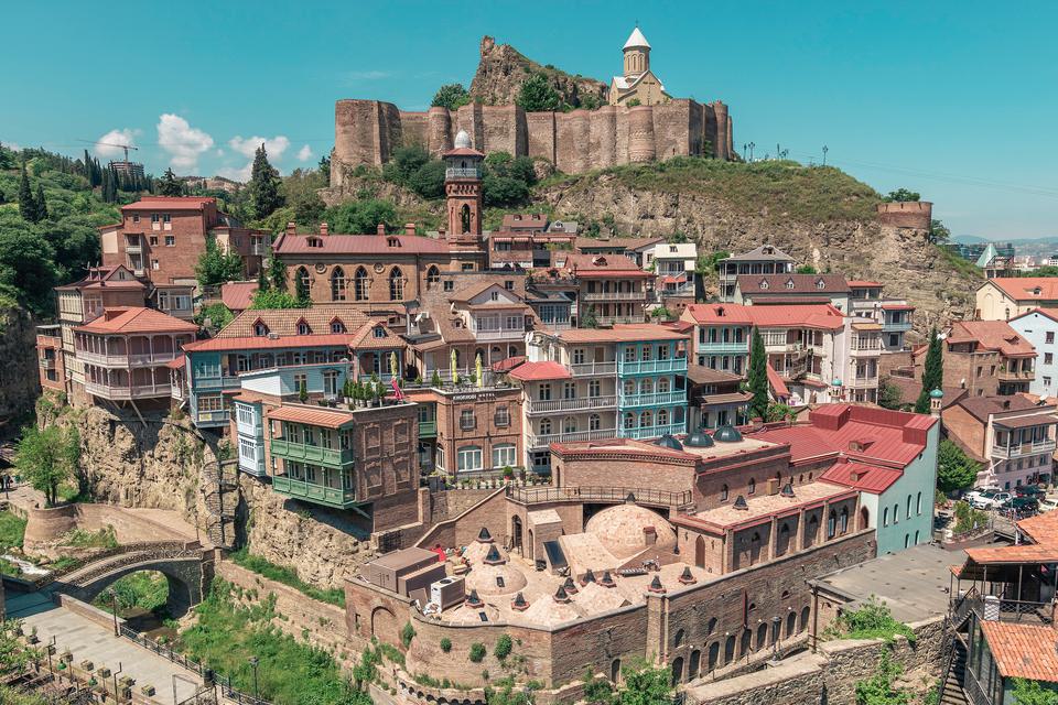 Old Tbilisi on a clear day: houses with carved wooden balconies and the brick domes of the sulphur baths climb the hillside below Narikala Fortress and St Nicholas Church.